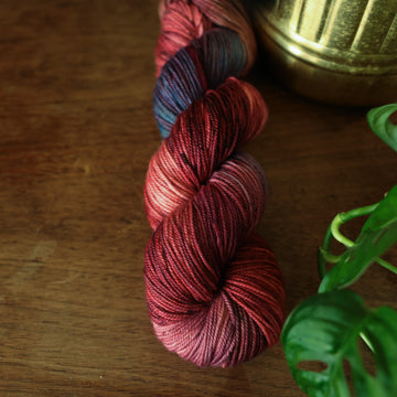 Sport yarn colored with different shades of red and some purple and blue colors mixed in, on top of a brown wooden table and a golden pot in the corner with a plant peaking out.