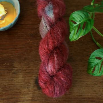 Suri alpaca yarn colored with different shades of red and some purple and blue colors mixed in, on top of a brown wooden and a partially melted candle in the corner with a plant peaking out.