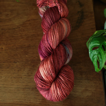 Worsted yarn colored with different shades of red and some purple and blue colors mixed in, on top of a brown wooden table with a plant peaking out.