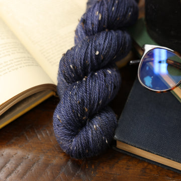 Dark blue yarn with brown black and white speckles on it, with an open book underneath it and two closed books next to it and a pair of glasses on top, all on top of a dark wooden table.