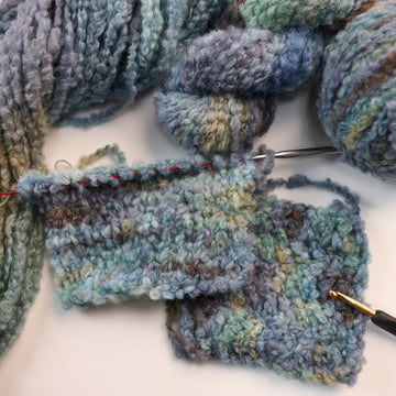 A dark purple/blue, light teal/green, and dark grey colored fluffy yarn on top of un unwound version of that yarn, and a knit swatch and crochet swatch with knitting needles and a crochet hook next to the yarn.