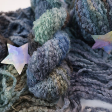A dark purple/blue, light teal/green, and dark grey colored fluffy yarn on top of un unwound version of that yarn, and two multicolored stars next to the yarn.