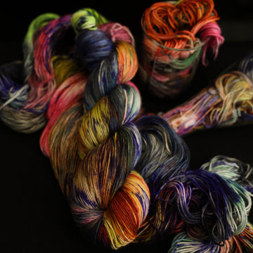 Multi colored fingering-3 yarn on top with more unwound multi colored yarn underneath and in a science beaker all on top of a black table
