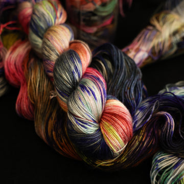 Multi colored silk square yarn on top with more unwound multi colored yarn underneath and in a science beaker all on top of a black table