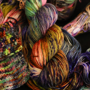 Multi colored DK yarn on top with more unwound multi colored yarn underneath and in a science beaker all on top of a black table and swatches on the side.