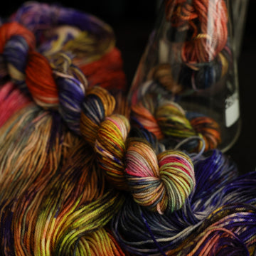 Multi colored fingering mini yarn on top with more unwound multi colored yarn underneath and another mini in a science beaker, all on top of a black table