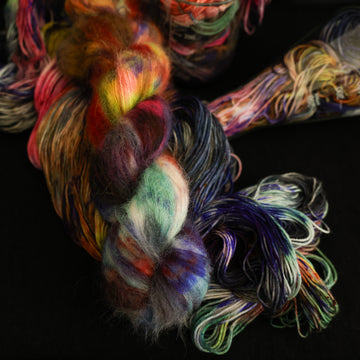 Multi colored suri yarn on top with more unwound multi colored yarn underneath and in a science beaker all on top of a black table