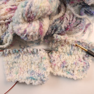 A fluffy white yarn with colors of light pink, purple, and blue, on top of an unwound version of that yarn, with some crochet and knit swatches of that yarn next to them, with a crochet needle and knitting needles next to it.