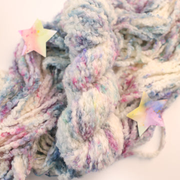 A fluffy white yarn with colors of light pink, purple, and blue, on top of an unwound version of that yarn, on a white table, with some multicolored stars around the yarn.