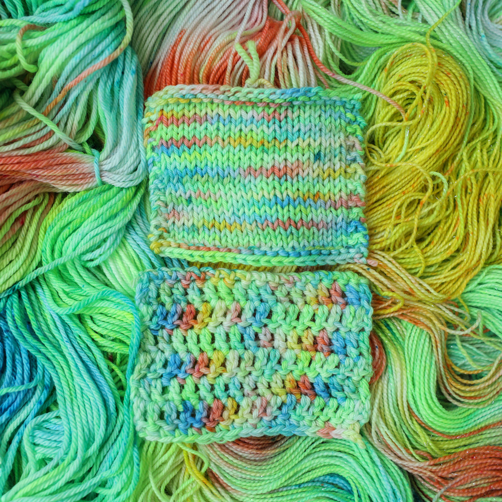 crochet and knit swatches colored different colors like a light neon green, light blue, some streaks of white, some red, and some yellow. These swatches are on top of unwound skeins of the same color way.