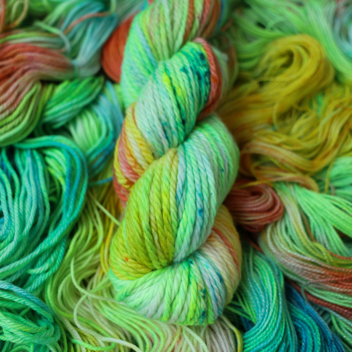 Bulky yarn colored different colors like a light neon green, light blue, some streaks of white, some red, and some yellow. This skein is on top of unwound skeins of the same color way.