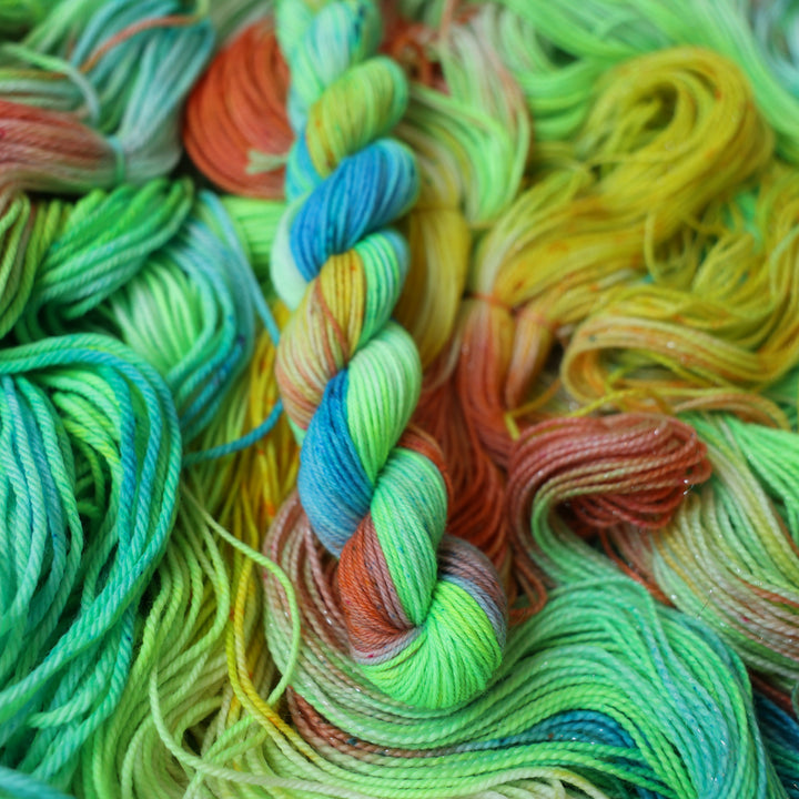 Fingering mini yarn colored different colors like a light neon green, light blue, some streaks of white, some red, and some yellow. This skein is on top of unwound skeins of the same color way.