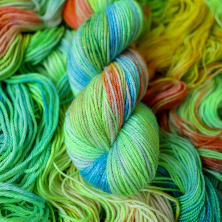 Sparkle DK yarn colored different colors like a light neon green, light blue, some streaks of white, some red, and some yellow. This skein is on top of unwound skeins of the same color way.