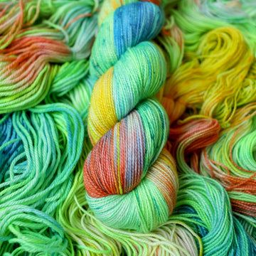 Sparkle fingering yarn colored different colors like a light neon green, light blue, some streaks of white, some red, and some yellow. This skein is on top of unwound skeins of the same color way.
