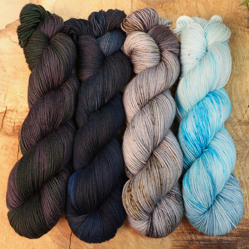 Nightfrost yarn set on a wooden table, the skiens from left to right are black and green, black and dark blue, grey with colored speckles, and icy blue.