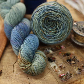A light grey/blue and light green worsted yarn next to some fishing equipment like hooks and ends of a pole. and a camping mug in the back along with a caked version of the yarn. all on top of a wooden table.