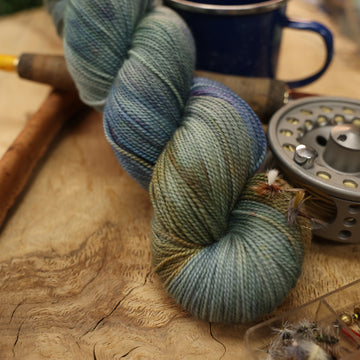 A light grey/blue and light green yarn next to some fishing equipment like hooks and ends of a pole. and a camping mug in the back, all on top of a wooden table.