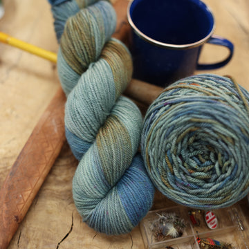 A light grey/blue and light green DK yarn next to some fishing equipment like hooks and ends of a pole. and a camping mug in the back along with a caked version of the yarn. all on top of a wooden table.