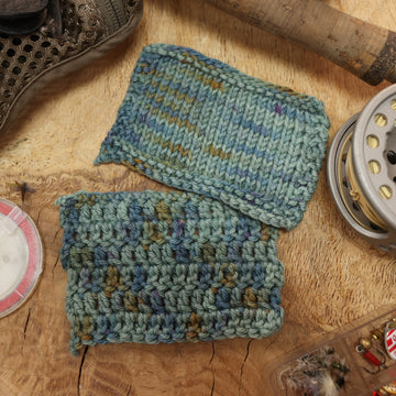 A light grey/blue and light green knit and crochet swatches next to some fishing equipment like hooks, the end of a pole, and fishing wire, all on top of a wooden table.