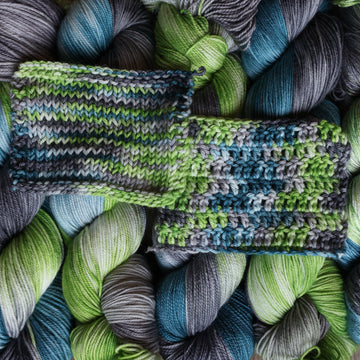 Wound skeins stacked next to each other that are Light lime green, Blue, and grey, colored with some white mixed in, with the same yarn that is in a knit and crochet swatch on top.