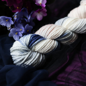 A cream colored Bulky skein with a streak of dark blue and hues of light pink going through the cream color. This skein is on top of a dark purple tablecloth and dark blackish blue tablecloth with blue and purplish pink flowers laying next to the skein on both sides.