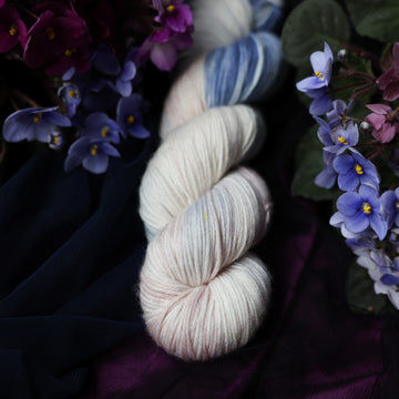 A cream colored DK skein with a streak of dark blue and hues of light pink going through the cream color. This skein is on top of a dark purple tablecloth and dark blackish blue tablecloth with blue and purplish pink flowers laying next to the skein on both sides.