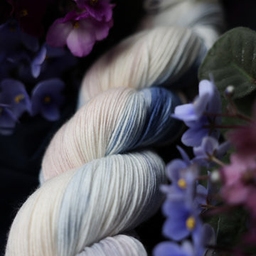 A cream colored skein with a streak of dark blue and hues of light pink going through the cream color. This skein is on top of a dark purple tablecloth and dark blackish blue tablecloth with blue and purplish pink flowers laying next to the skein on both sides. This is a close up picture with lots of details.