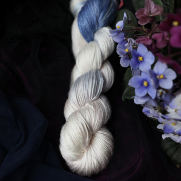 A cream colored Silk skein with a streak of dark blue and hues of light pink going through the cream color. This skein is on top of a dark purple tablecloth and dark blackish blue tablecloth with blue and purplish pink flowers laying next to the skein on both sides.