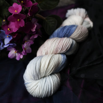 A cream colored Sparkle DK skein with a streak of dark blue and hues of light pink going through the cream color. This skein is on top of a dark purple tablecloth and dark blackish blue tablecloth with blue and purplish pink flowers laying next to the skein on both sides.