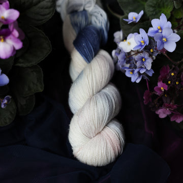 A cream colored Sparkle Fingering skein with a streak of dark blue and hues of very light pink going through the cream color. This skein is on top of a dark purple tablecloth and dark blackish blue tablecloth with blue and purplish pink flowers laying next to the skein on both sides.