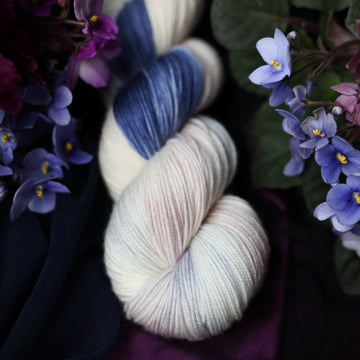 A cream colored Sport skein with a streak of dark blue and hues of light pink going through the cream color. This skein is on top of a dark purple tablecloth and dark blackish blue tablecloth with blue and purplish pink flowers laying next to the skein on both sides.