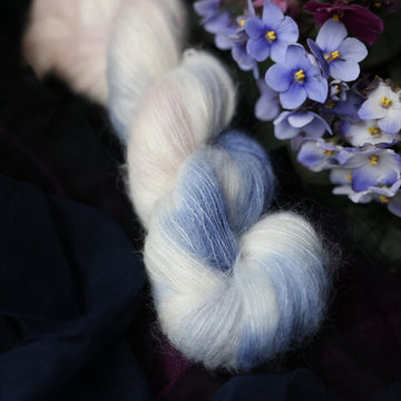 A cream colored Suri skein with a streak of dark blue and hues of light pink going through the cream color. This skein is on top of a dark purple tablecloth and dark blackish blue tablecloth with blue and purplish pink flowers laying next to the skein on both sides.