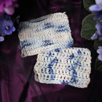 A cream colored crochet and knit swatches with streaks of dark blue and hues of light pink going through the cream color. This skein is on top of a dark purple tablecloth with blue and purplish pink flowers laying next to the skein on both sides.