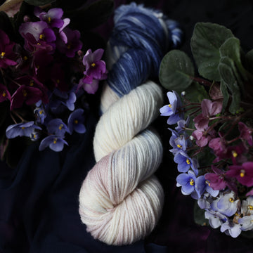 A cream colored Worsted skein with a streak of dark blue and hues of light pink going through the cream color. This skein is on top of a dark purple tablecloth and dark blackish blue tablecloth with blue and purplish pink flowers laying next to the skein on both sides.