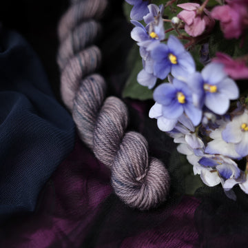 A DK Mini yarn colored  romantic plum-grey with moody grey and blue coloring. Surrounded by colorful flowers all sitting on top of dark blue and purple fabric.