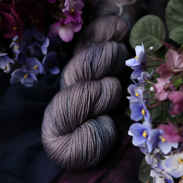 A fingering skeined yarn colored  romantic plum-grey with moody grey and blue coloring. Surrounded by colorful flowers all sitting on top of dark blue and purple fabric.