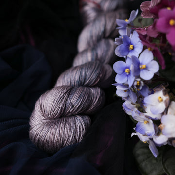 A silk skein yarn colored  romantic plum-grey with moody grey and blue coloring. Surrounded by colorful flowers all sitting on top of dark blue fabric.