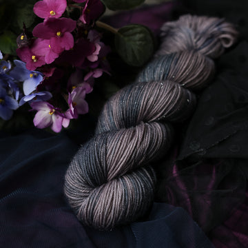 A sparkle DK skein yarn colored  romantic plum-grey with moody grey and blue coloring. Surrounded by colorful flowers all sitting on top of dark blue fabric.