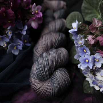A Sport skein yarn colored  romantic plum-grey with moody grey and blue coloring. Surrounded by colorful flowers all sitting on top of dark blue and purple fabric.
