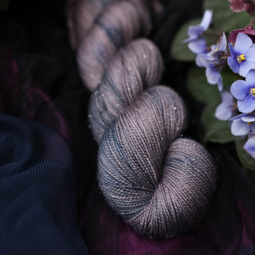 A sparkle fingering skeined yarn colored  romantic plum-grey with moody grey and blue coloring. Surrounded by colorful flowers all sitting on top of dark blue and purple fabric.