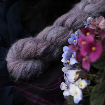 A Suri skein yarn colored  romantic plum-grey with moody grey and blue coloring. Surrounded by colorful flowers all sitting on top of dark blue and purple fabric.