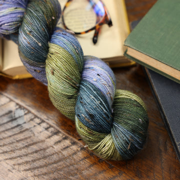 different colored blue green and white yarn on top of a wooden table with books next to it and a pair of glasses in the top of the picture.