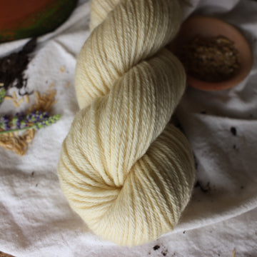 A light cream colored Worsted yarn skein on top of a white tablecloth with some flowers and rice grains and a plant pot next to it.