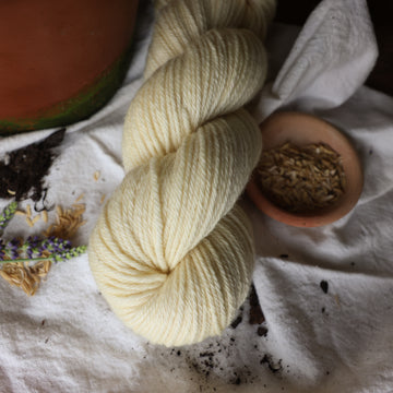 A light cream colored Dk yarn skein on top of a white tablecloth with some flowers and rice grains and a plant pot next to it.