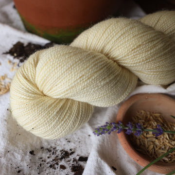 A light cream colored Fingering yarn skein on top of a white tablecloth with some flowers and rice grains and a plant pot next to it.
