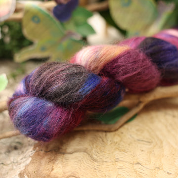 a dark purple, pink, and red colored soft alpaca yarn with some speckles on it, on top of a plant with bare branches and leaves in the back. along with painted butterflies and leaves made of book pages, all on top of a wooden table.