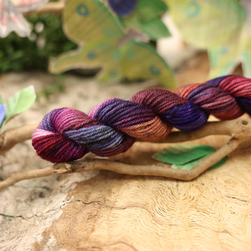 a dark purple, pink, and red colored DK mini yarn with some speckles on it, on top of a plant with bare branches and leaves in the back. along with painted butterflies and leaves made of book pages, all on top of a wooden table.