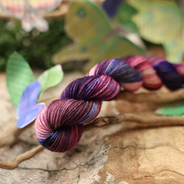 a dark purple, pink, and red colored fingering mini yarn with some speckles on it, on top of a plant with bare branches and leaves in the back. along with painted butterflies and leaves made of book pages, all on top of a wooden table.