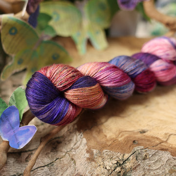 a dark purple, pink, and red colored silk yarn with some speckles on it, on top of a plant with bare branches and leaves in the back. along with painted butterflies and leaves made of book pages, all on top of a wooden table.
