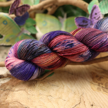a dark purple, pink, and red colored sparkle yarn with some speckles on it, on top of a plant with bare branches and leaves in the back. along with painted butterflies and leaves made of book pages, all on top of a wooden table.